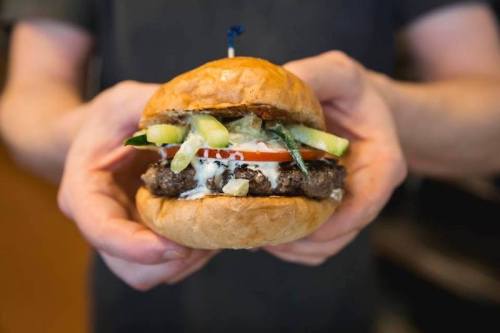 A person holding a juicy burger with lettuce, tomato, pickles, and sauce, topped with a blue toothpick.