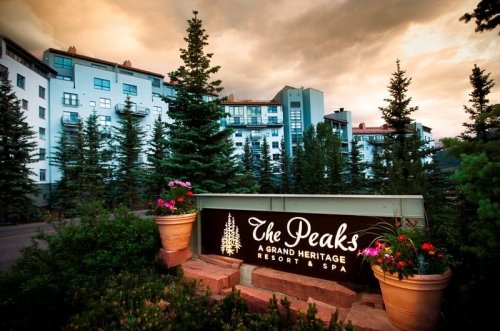 Sign for The Peaks Resort & Spa surrounded by trees and flowers, with a scenic backdrop of a cloudy sky.