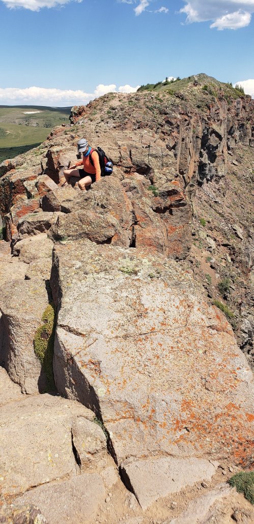A person crouches on a rocky cliff edge, surrounded by expansive green hills and a blue sky with clouds.