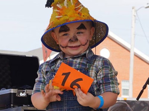A smiling child in a scarecrow costume holds a number 16 sign, with face paint and a colorful hat.