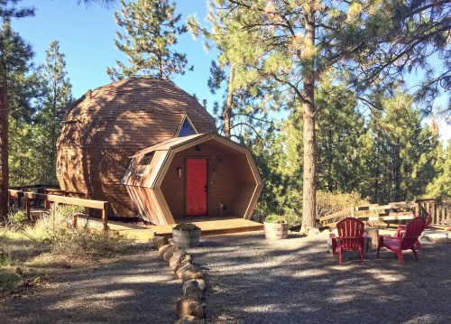 A unique, modern cabin with a round structure and hexagonal extension, surrounded by trees and outdoor seating.