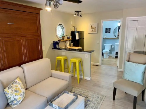 Cozy living space with a sofa, yellow bar stools, kitchenette, and a bathroom visible in the background.