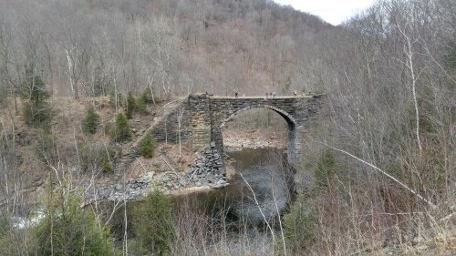 A stone arch bridge spans a river, surrounded by bare trees and rocky terrain in a mountainous area.