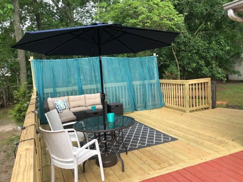 A wooden deck with a seating area, umbrella, and sheer curtains, surrounded by greenery.