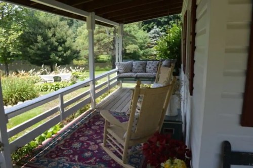 A cozy porch with rocking chairs, a patterned rug, and a view of a lush garden and trees in the background.
