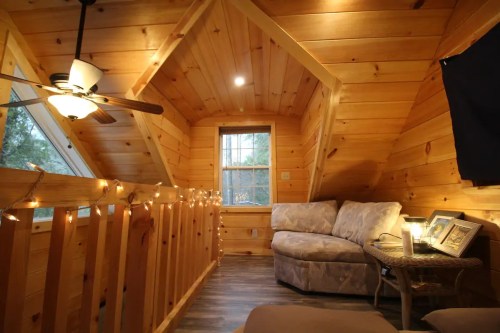 Cozy wooden loft with a couch, ceiling fan, and string lights, featuring large windows and a warm, inviting atmosphere.