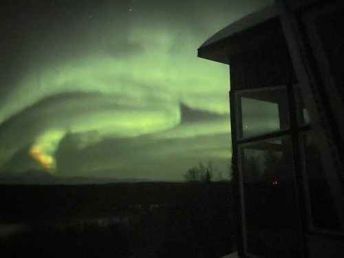 A view of swirling green auroras in the night sky, with a building silhouette in the foreground.