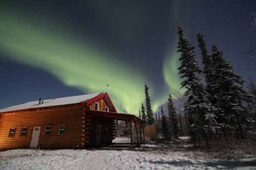 A log cabin under a vibrant green aurora borealis, surrounded by snow and tall trees at night.