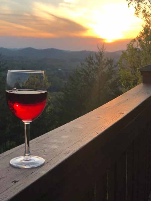 A glass of red wine sits on a wooden railing, with a sunset over rolling hills in the background.