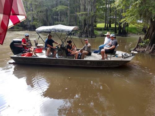 A group of six people sits on a boat in a swampy area, surrounded by trees and water.