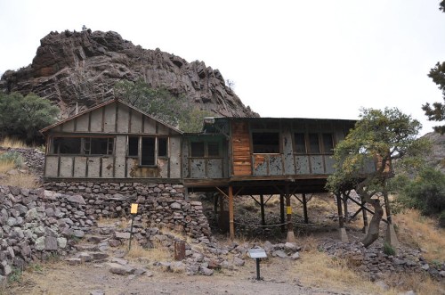 A rustic, two-story cabin with a stone foundation, set against a rocky hillside and surrounded by sparse vegetation.