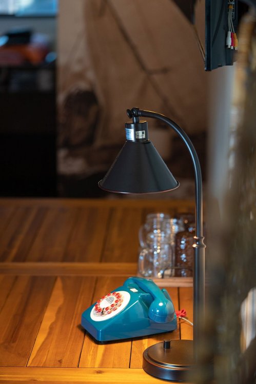 A blue rotary phone sits on a wooden table beside a black lamp, with glassware in the background.