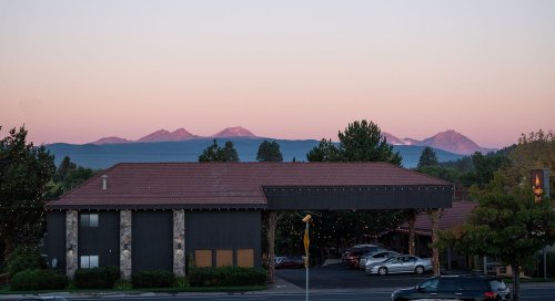 A view of mountains at sunset, with a building in the foreground and a clear sky transitioning to pink hues.