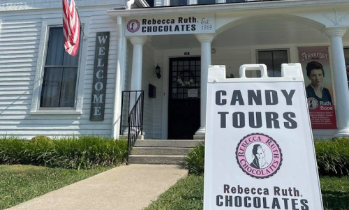 A white building with a sign for Rebecca Ruth Chocolates and a sandwich board advertising candy tours.