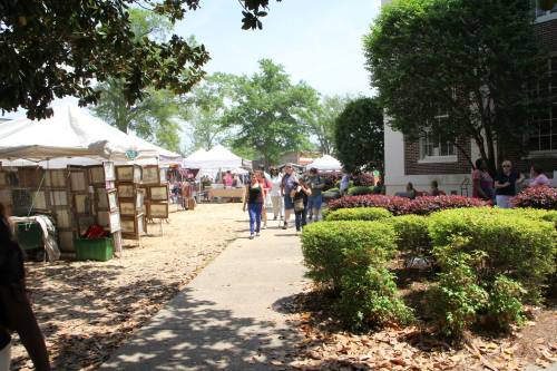 A bustling outdoor market with tents, people walking, and greenery lining the pathway on a sunny day.