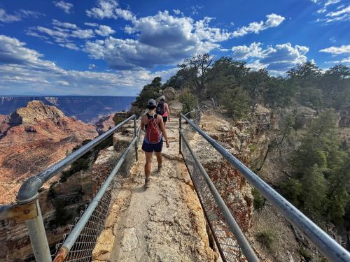 Two hikers walk along a narrow path with a railing, overlooking a vast canyon under a blue sky with scattered clouds.