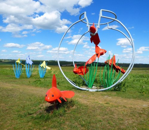 Colorful metal sculptures of goldfish and flowers against a blue sky with fluffy clouds in a grassy landscape.