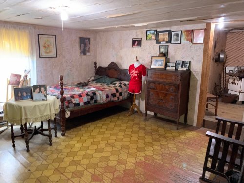 A cozy, vintage bedroom with a quilted bed, a wooden dresser, framed photos, and a mannequin wearing a red shirt.