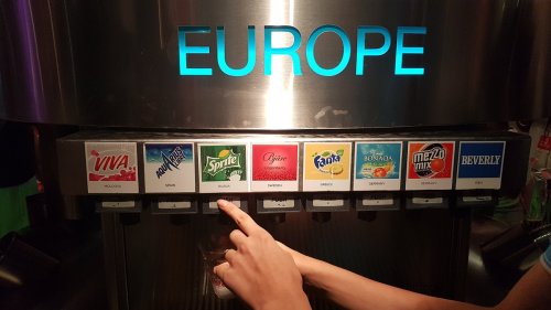A beverage dispenser labeled "EUROPE" with various soda options and a hand selecting a drink.
