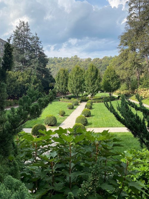 A lush garden with neatly trimmed hedges, pathways, and trees under a partly cloudy sky.