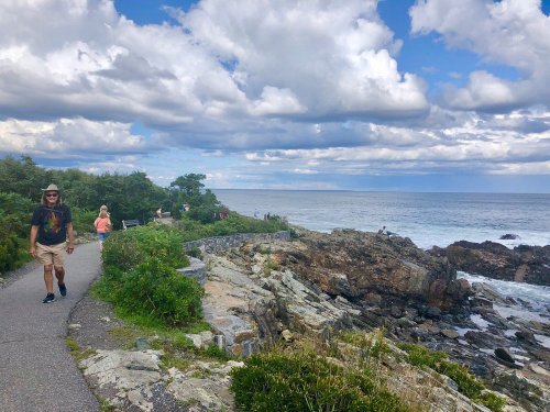 A scenic coastal path with rocky shores, greenery, and people walking under a partly cloudy sky.