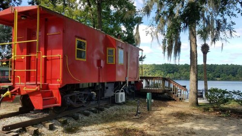 A bright red train caboose beside a wooden deck, surrounded by trees and overlooking a calm lake.