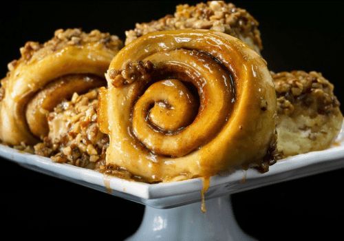 A close-up of sticky cinnamon rolls topped with nuts, displayed on a white plate against a dark background.