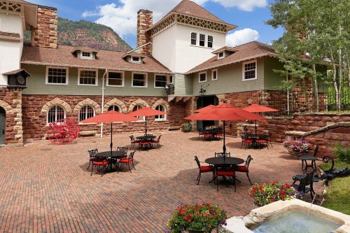 A charming outdoor patio with red umbrellas over black tables, surrounded by stone buildings and mountains in the background.