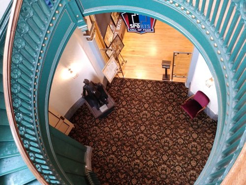 Aerial view of a staircase leading to a carpeted hall with a statue and a chair, framed by ornate railings.