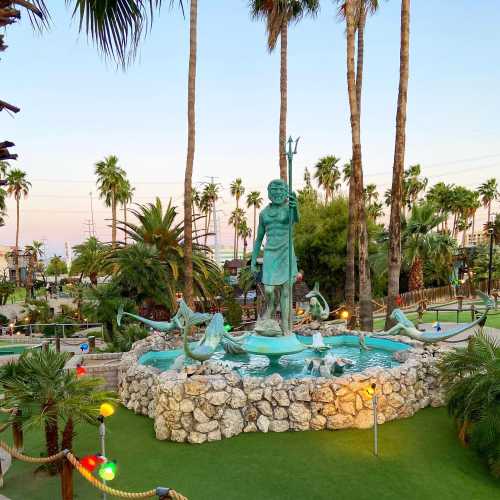 A statue surrounded by palm trees and colorful lights, set in a park with a fountain and greenery at sunset.