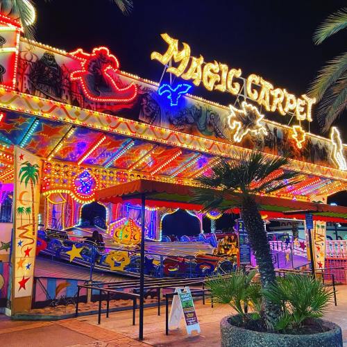 Brightly lit carnival ride named "Magic Carpet" with colorful lights and palm trees in the foreground at night.