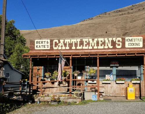 A rustic restaurant with a wooden facade, flower pots, and an American flag, set against a hilly landscape.