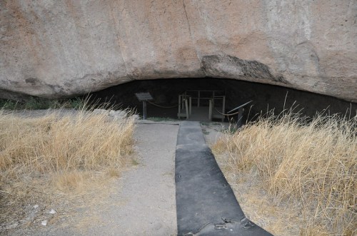 Entrance to a cave under a large rock formation, with dry grass and a pathway leading inside.