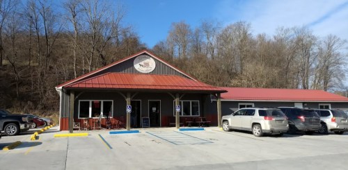 A rustic building with a red roof, surrounded by trees, featuring parked cars in front and a sign above the entrance.