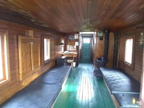 Interior of a wooden cabin with two beds, a table, and a green door, featuring natural light from windows.