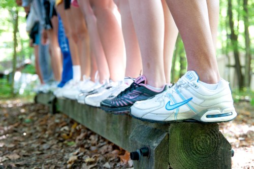 A row of feet in various athletic shoes balanced on a wooden beam in a forested area.