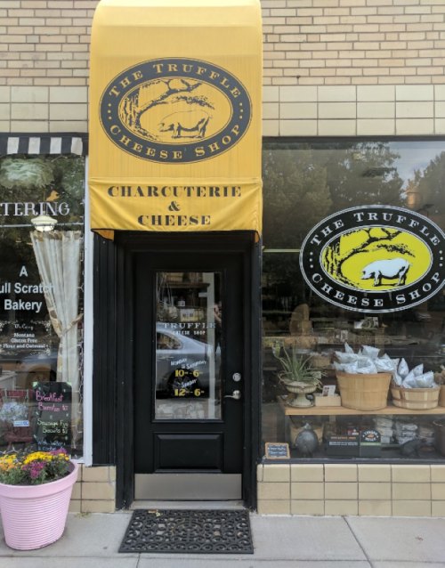 Entrance of "The Truffle Cheese Shop," featuring a yellow awning and a display of cheese and charcuterie products.
