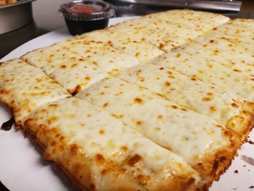 A close-up of a cheesy pizza with golden crust, cut into squares, served with a small container of marinara sauce.