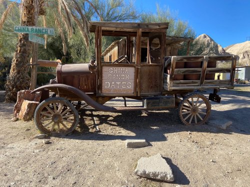 An old, weathered truck with "China Ranch Dates" signage, parked near a palm tree and a road sign.