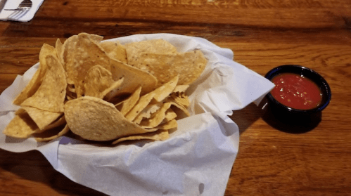 A basket of tortilla chips on a wooden table, accompanied by a small bowl of red salsa.
