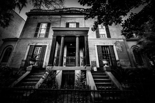 A historic, two-story house with a grand entrance, surrounded by lush greenery, captured in black and white.