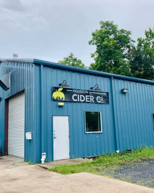 Exterior of a blue metal building with a sign that reads "Houston Cider Co" and a white door. Green trees in the background.