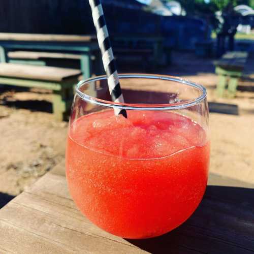 A refreshing red slush drink in a glass with a black and white striped straw, set on a wooden table outdoors.