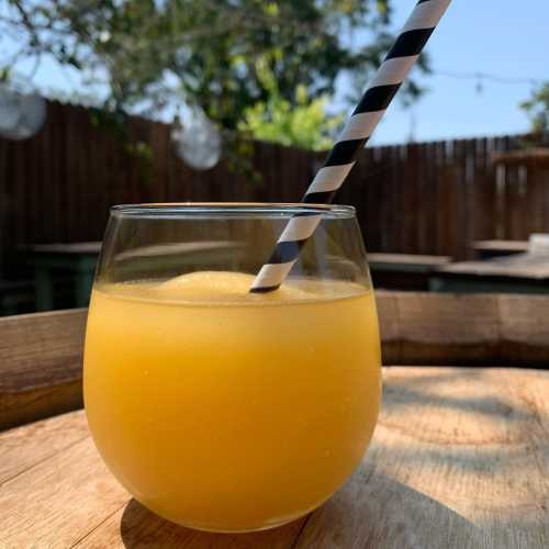 A glass of orange smoothie with a black and white striped straw, set on a wooden table outdoors.