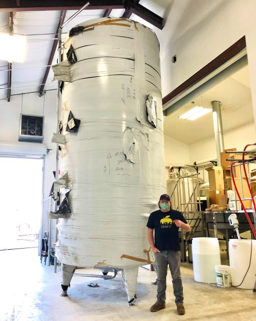 A person stands next to a large, wrapped cylindrical tank in an industrial setting, wearing a mask and holding a tool.