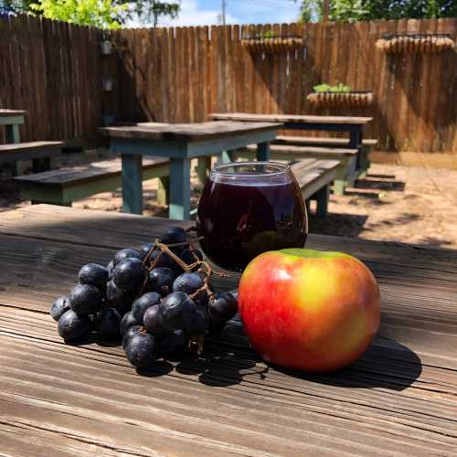 A red apple and a bunch of black grapes beside a glass of dark liquid on a wooden table in an outdoor setting.