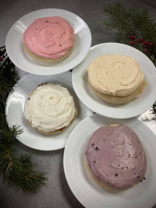 Four round cakes with different colored frostings on white plates, surrounded by pine branches and festive decorations.