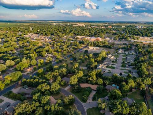 Aerial view of a suburban neighborhood with green trees, houses, and open fields under a partly cloudy sky.