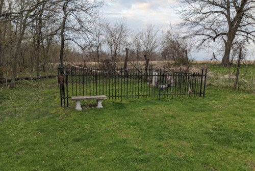 A small fenced area with a stone bench, surrounded by grass and trees, under a cloudy sky.