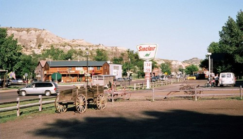 A small town scene featuring a Sinclair gas station, wooden buildings, and a wagon in the foreground with hills in the background.